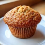 A close-up shot of one perfectly baked, moist Banana Muffins with a golden-brown, slightly glossy top, resting on a white plate.