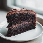 Close-up of a moist, dark slice of Chocolate Fudge Cake with rich chocolate frosting on a white plate.