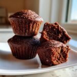 Three dark brown Gingerbread Muffins stacked on a white plate, one broken open to show the moist crumb.