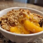 Close-up of a white bowl filled with warm Not Your Average Peach Crisp, showing juicy peaches and crumb topping.