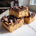 Three stacked Peanut Butter Cookie Dough Bars topped with melted chocolate and chocolate chips on a white marble surface.