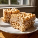Three squares of chewy Peanut Butter Rice Krispies treats stacked slightly on a white plate near a window.