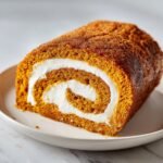 A close-up of a slice of moist Pumpkin Roll Cake featuring an orange sponge rolled tightly with white cream cheese filling.