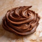A close-up swirl of rich, dark brown Chocolate Frosting sitting on a light marble surface.