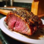 Close-up of a medium-rare Roast Leg Of Lamb slice seasoned with herbs on a white plate.