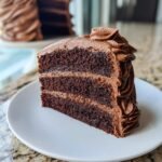 A generous slice of three-layer Chocolate Buttercream Cake with rich frosting layers shown on a white plate.