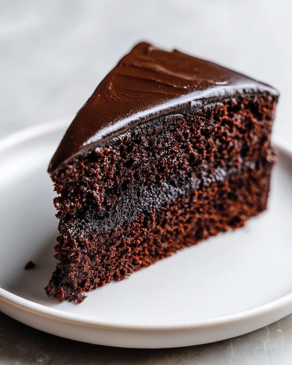 A close-up of a moist slice of dark Chocolate Cake topped with smooth, glossy ganache frosting on a white plate.