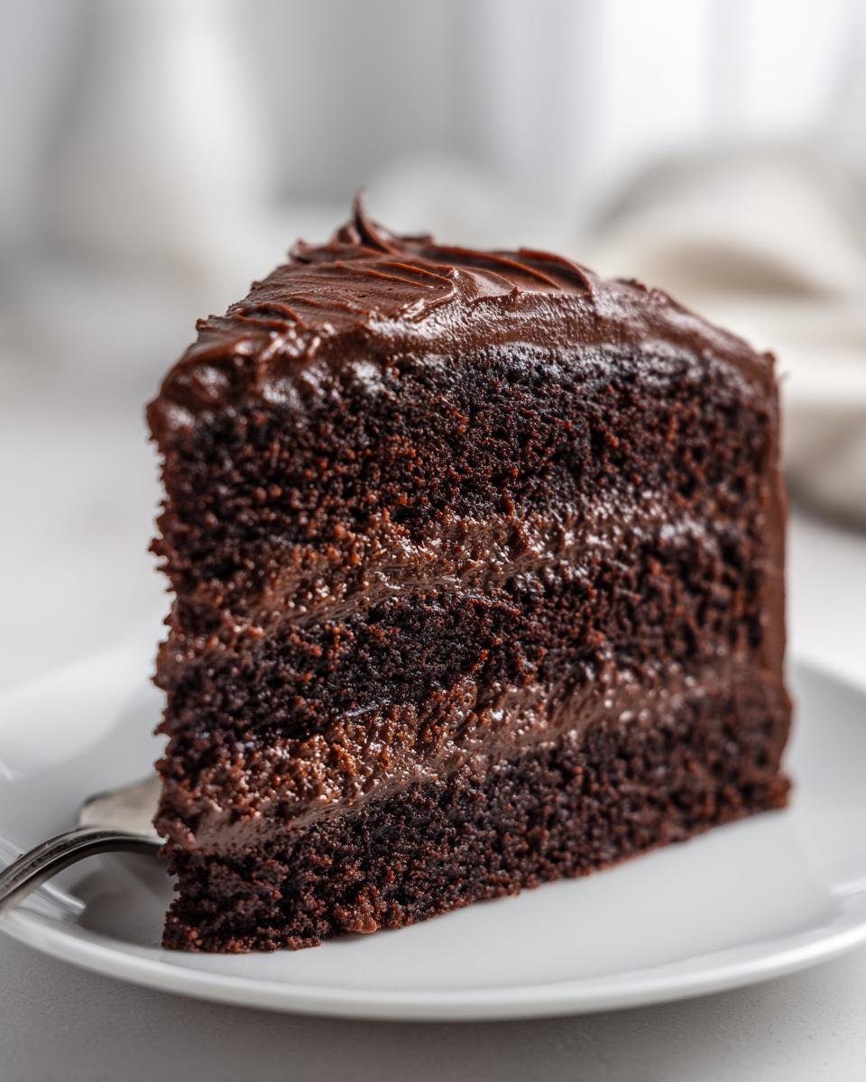 A decadent slice of moist Chocolate Fudge Cake layered with thick fudge frosting on a white plate.