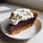 A rich slice of Chocolate Pudding Pie with a graham cracker crust, topped generously with whipped cream.