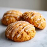 Three soft, orange Pumpkin Cookies drizzled with white vanilla icing, resting on a white marble surface.