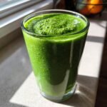 A tall glass filled with a thick, vibrant Super Green Detox Smoothie, sitting on a counter in bright sunlight.