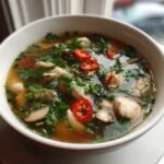A close-up of a white bowl filled with flavorful Thai Chicken Soup, featuring chicken pieces, mushrooms, fresh cilantro, and red chili slices.