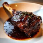 A perfectly cooked Wine Braised Lamb Shank glazed in a dark sauce and topped with fresh rosemary sprigs.