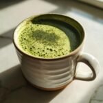 A close-up of a vibrant green, foamy matcha hot chocolate in a white, textured mug.