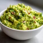 Close-up of a white bowl filled with The Best Guacamole Recipe Creamy Flavorful Delight, showing chunks of avocado, onion, and cilantro.