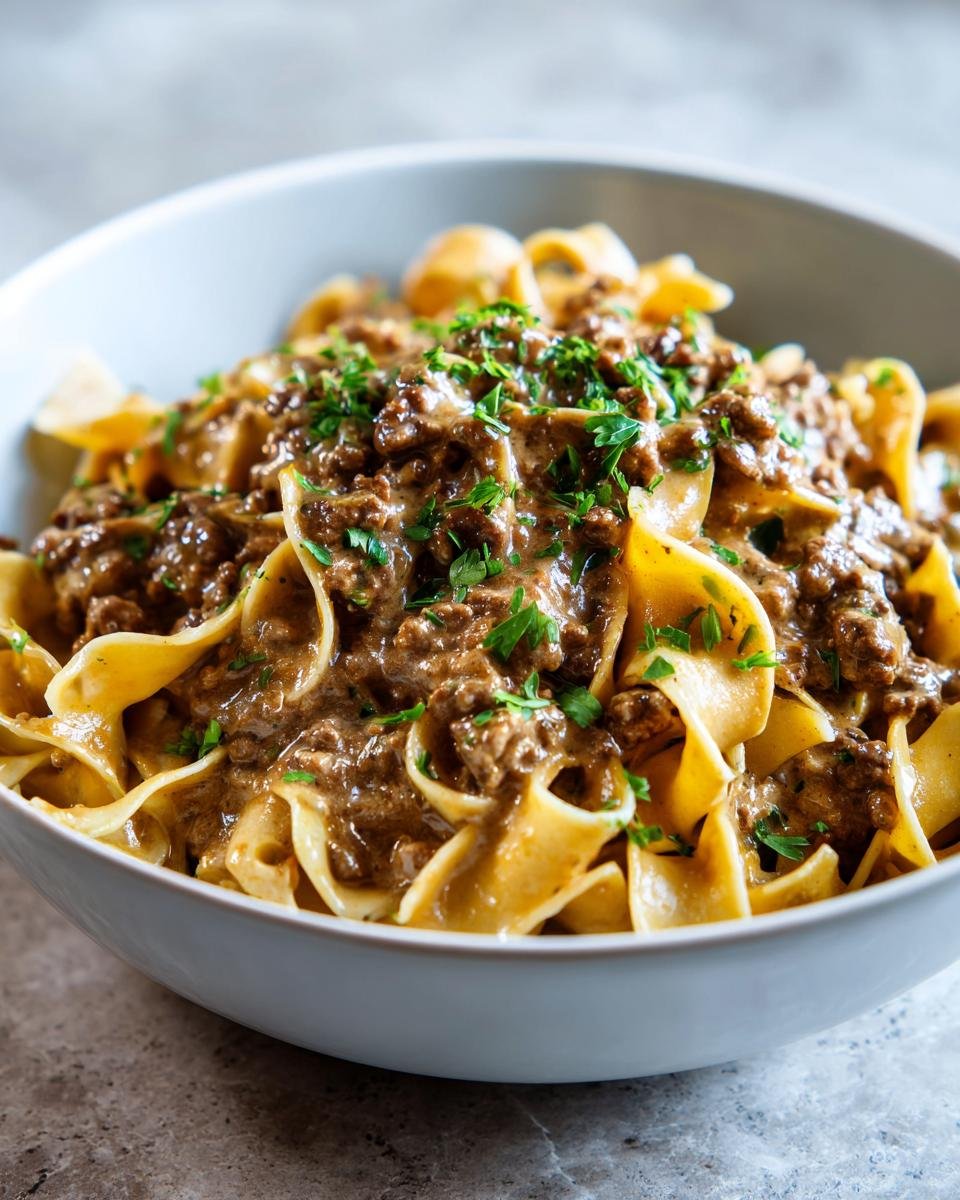 A close-up of a bowl filled with Cozy Beef And Noodles, featuring wide egg noodles coated in a rich, creamy beef sauce and topped with fresh parsley.