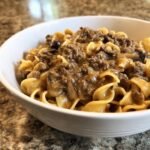 A close-up of a white bowl filled with Cozy Beef And Noodles, featuring egg noodles and a rich, savory ground beef and mushroom sauce.