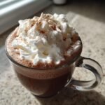 A clear glass mug filled with Cozy Cinnamon Hot Chocolate topped high with whipped cream and a dusting of cinnamon.