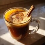 A close-up of a Cozy Hot Buttered Rum Recipe served in a clear glass mug, topped with foam and a cinnamon stick.
