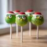 Several Delightful Grinch Cake Pops decorated with green coating, Santa hats, and candy eyes, sitting on a wooden surface.