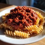 A close-up of Hearty Lentil Bolognese Comfort Food served over a bed of spiral fusilli pasta on a white plate.