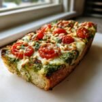 Close-up of a thick slice of Heavenly Pesto Pizza topped with bright green pesto, melted mozzarella, roasted cherry tomatoes, and pine nuts.