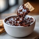 A cracker loaded with thick, glossy Indulgent Brownie Batter Dip being lifted from a white bowl.