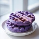A stack of two vibrant purple Irresistible Crispy And Tender Ube Waffles lightly dusted with powdered sugar on a white plate.