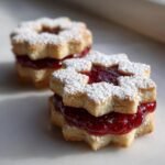 Two Irresistible Easy Linzer Cookies filled with bright red raspberry jam and dusted with powdered sugar.