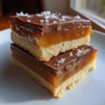 Two stacked squares of Irresistible Homemade Twix Bars recipe featuring shortbread, caramel, chocolate, and sea salt.