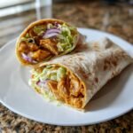 Two halves of Irresistible Honey Mustard Chicken Wraps filled with saucy chicken, lettuce, and red onion on a white plate.