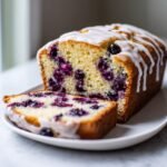 A loaf of Irresistible Lemon Blueberry Bread Recipe with a slice cut, topped with white lemon glaze.