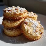 A stack of Irresistible Pignoli Cookies topped with sliced almonds and powdered sugar on a white plate.