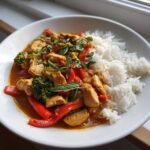 A bowl of Irresistible Red Curry Recipe featuring chicken, red peppers, and broccoli served next to fluffy white rice.