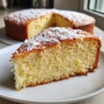 A close-up of a moist slice of Irresistible Yogurt Cake dusted with powdered sugar, with the rest of the cake in the background.
