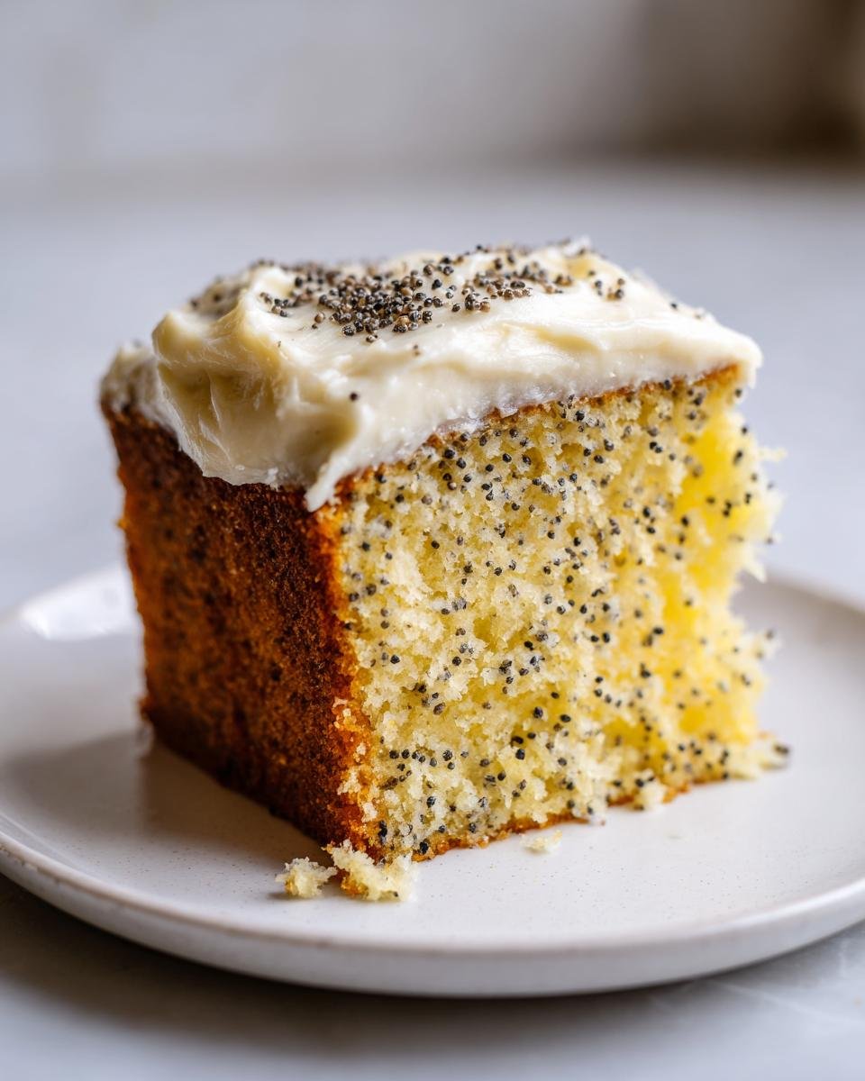 A close-up of a moist slice of Irresistible Lemon Poppy Seed Cake topped with thick cream cheese frosting and sprinkled poppy seeds.