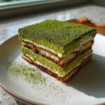 A perfect square slice of Matcha Tiramisu showing layers of ladyfingers and green cream, dusted heavily with matcha powder.