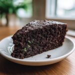 A moist slice of dark chocolate zucchini cake showing flecks of green zucchini, served on a white plate.