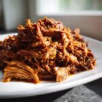 A close-up of shredded, saucy, Mouthwatering Pulled Pork Recipe piled high on a white plate.