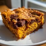 A close-up of a moist, orange-hued Paleo Pumpkin Blondies square topped with melted chocolate chunks on a white plate.