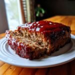 A close-up of a thick slice of Ultimate Moist Meatloaf Recipe covered in a glossy, red glaze.