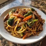 A close-up of a white plate filled with Irresistible Veggie Chicken Lo Mein Recipe, featuring noodles, broccoli, carrots, and chicken.