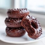 A stack of rich, dark Baked Chocolate Donuts Grain Free Paleo, with one donut bitten into, showing the moist interior.