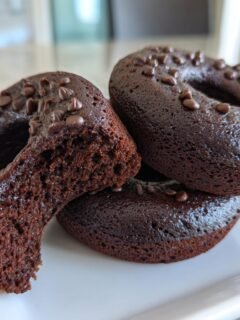 Three rich, dark Baked Chocolate Donuts Grain Free Paleo stacked on a white plate, one cut to show the moist interior.