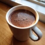Close-up of a steaming mug of rich Bone Broth Hot Chocolate with a bubbly top, set on a wooden surface near a window.