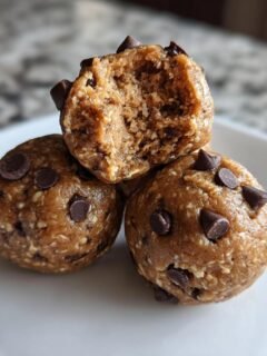 A stack of three chocolate chip Cookie Dough Protein Balls, one on top is broken open showing the texture.
