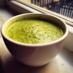 Close-up of vibrant green Creamy Broccoli Cheese Soup Paleo Whole30 Vegan served in a white bowl by a window.