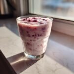A creamy Coconut Milk Smoothie with visible red berry pieces, served in a glass by a sunny window.