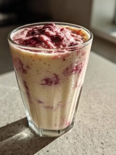 A tall glass of creamy Coconut Milk Smoothie swirled with vibrant red berries, sitting on a speckled countertop.