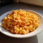 A close-up of creamy, orange High Protein Mac And Cheese served in a white bowl under bright sunlight.